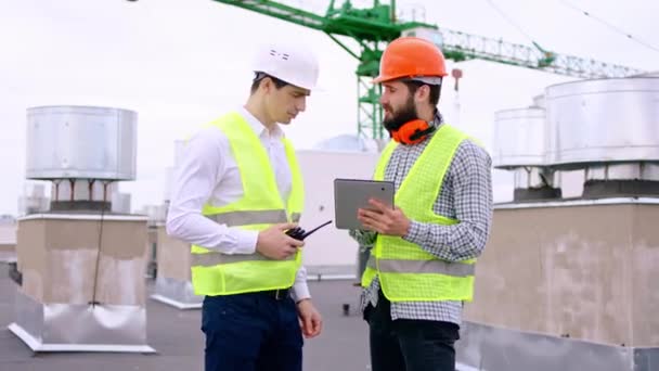 Attractive engineers at construction site with a safety helmets on the top of the building using digital tablet together discussing some details of the plan of construction. 4k — Stock Video