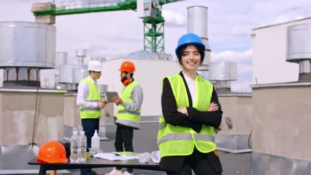 Bella assistente ingegnere donna guardando dritto alla fotocamera e sorridente grande mentre in piedi sul tetto del cantiere altro architetto e ingegnere sullo sfondo discutendo — Video Stock