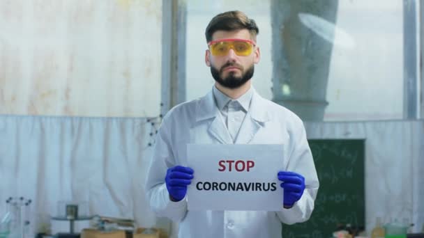 A biotechnológiai laborban a kameratudós ember előtt egy Stop Coronavirus poszterrel a kezében egyenesen a COVID-19 kamera koncepciójára nézett. — Stock videók