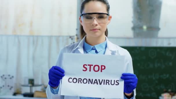 화학 실험실에 있는 아름다운 여성 과학자가 전 세계적으로 유행하고 있는 Stop Coronavirus 의 포스터를 들고 있는 모습을 카메라 앞에 클로즈업하고 있습니다. 4k — 비디오