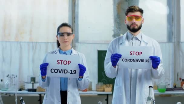 Dos científicos y un hombre sosteniendo dos carteles de Stop Coronavirus en el laboratorio de química miran directamente a la cámara — Vídeos de Stock