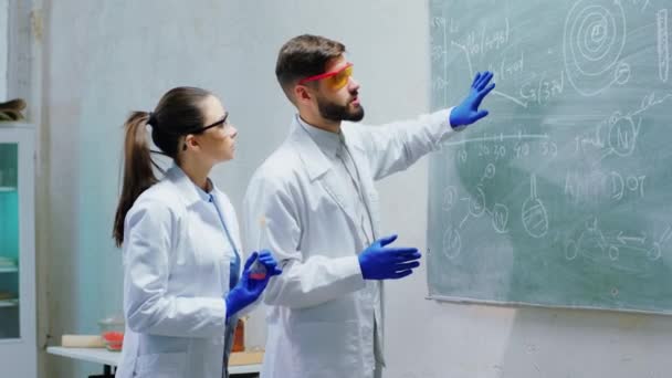 W laboratorium chemicznym naukowiec i jego asystent razem przeprowadzają eksperyment, analizują notatki z tablicy i rozmawiają o podekscytowaniu. — Wideo stockowe