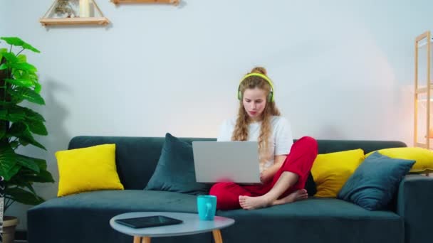 Молода студентка робить свій курс онлайн на ноутбуці, сидячи на дивані і слухаючи музику одночасно з навушниками — стокове відео