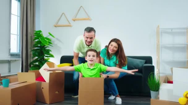 Em uma bela casa nova pais felizes e animados empurrando seu filho enquanto ele está na caixa na nova espaçosa sala de estar — Vídeo de Stock