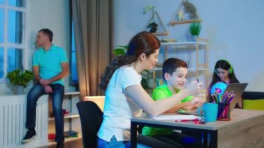 Modern bir oturma odasında, diğer aile üyeleri kanepede ya da pencerede vakit geçirirken küçük oğluyla ev ödevlerini yapıyor. 4k
