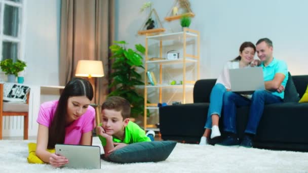 Geniş oturma odasında ebeveynler ve çocukları bilgisayar kullanarak vakit geçirmek için aletler kullanıyorlar ve çocuklar dijital tabletle oynuyorlar. — Stok video