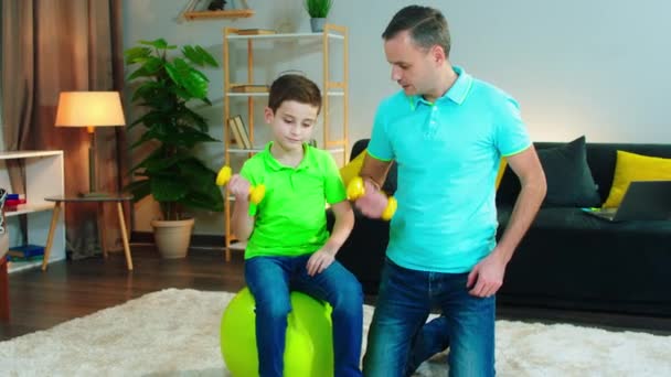 Tiempo de entrenamiento para papá y su lindo hijo que juntos haciendo ejercicios para hacer músculo usando los pesos en la sala de estar — Vídeos de Stock