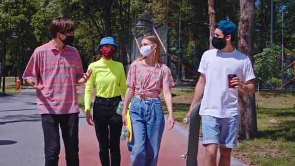 現代のスケートパークでのコロナウイルスのパンデミックで友人の男と女性のスタイリッシュなグループは、彼らが歩くと彼らのスケートボードの男の1を保持保護マスクを着て横にスケートボード — ストック動画