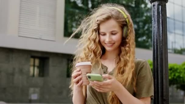 Добре виглядає молода жінка з довгим кучерявим волоссям посеред вуличної розмови з кимось на її смартфоні і тримає чашку кави. 4k — стокове відео