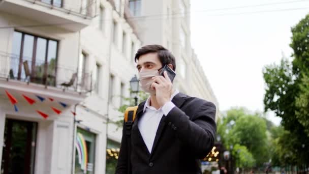 Charismatische zakenman met een beschermend masker die op straat loopt en op zijn smartphone voor de camera spreekt — Stockvideo