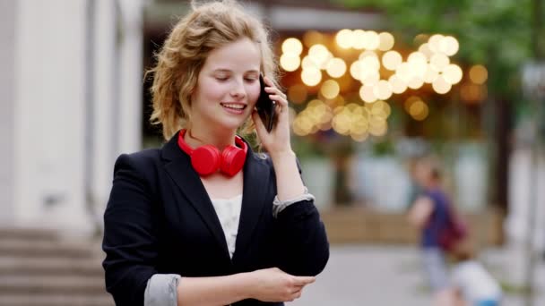Mujer bonita pelo rubio tener una conversación feliz y emocionada en su teléfono inteligente delante de la cámara en medio de la calle — Vídeos de Stock