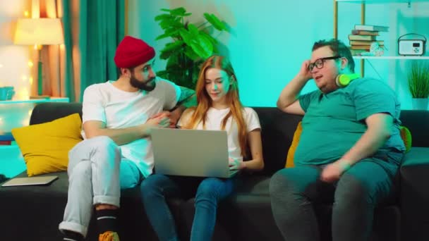 Enthousiaste et beau groupe d'amis deux gars et une jolie dame sur le canapé à la maison en utilisant un ordinateur portable ils regardent un film drôle — Video