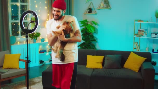 덩치큰 카리스마있는 남자가 귀여운 개와 노는 것에 영향을 주면서 거실에서 즐거운 시간을 보내는 그의 소셜 미디어 계정을 위해 라이브 스트림을 만들고 있습니다. — 비디오
