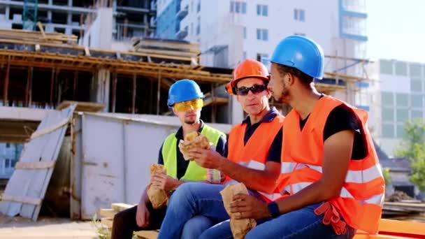 На обід у милі великого будівельного майданчика багатоетнічні робітники в захисних касках і окулярах їдять бутерброди і обговорюють — стокове відео