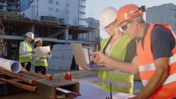 Charyzmatyczna młoda inżynier kobieta i brygadzista w średnim wieku analizując plan budowy razem za pomocą laptopa sprawdzają dokładnie noszenie munduru ochronnego i gogli — Wideo stockowe