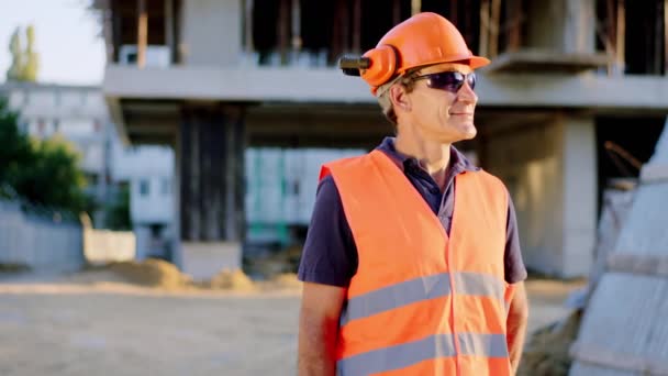 Před foťákem předák středního věku s bezpečnostní helmou a brýlemi ukazuje velké jako na konstrukci a usmívá pěkně — Stock video