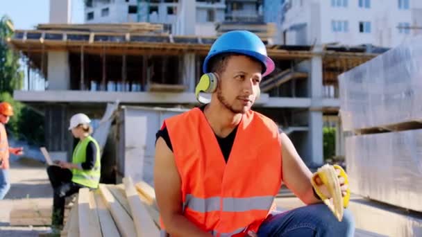 Молодий будівельник Афроамериканець під час перерви їсть банан і слухає музику перед камерою на будівельному майданчику. — стокове відео