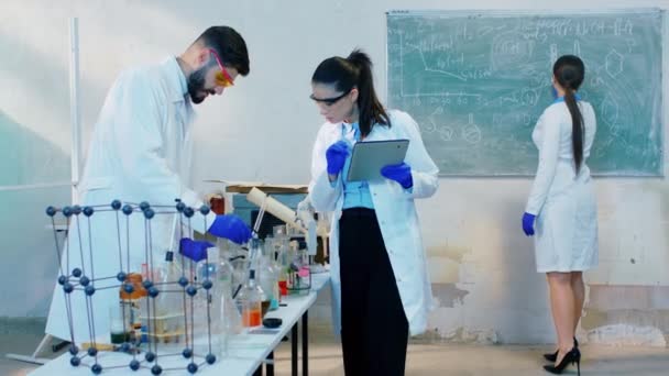 Los colegas científicos trabajando juntos en un experimento mezclaron la sustancia química y escribieron en la tableta digital los resultados que otra señora escribió alguna fórmula en la pizarra. Disparo en ARRI Alexa — Vídeo de stock