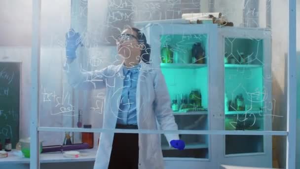 Izgalmas és koncentrált dolgozó nő tudós az átlátszó fórumon írja meg a kémiai képletet a kémiai laboratóriumában ő visel biztonsági felszerelést. Lelőtték az ARRI Alexa Mini-n. — Stock videók