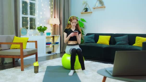 Una joven mujer sana está sentada en una gran bola de yoga amarilla, ella está en ropa de entrenamiento y se distrae con su teléfono también escuchando música — Vídeos de Stock