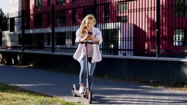 Attraktive Frau an einem sonnigen Tag mitten auf der Straße macht Halt mit ihrem Elektroroller und nutzt das Smartphone, um etwas zu sehen, das sie mit gelben Kopfhörern trägt — Stockvideo