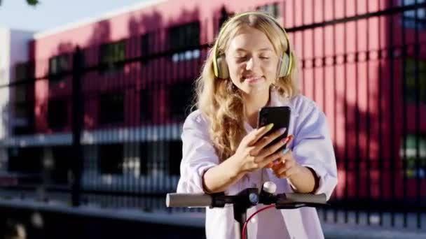 Κοντινό πορτρέτο του καταπληκτική όμορφη μαθήτρια στη μέση ενός ηλιόλουστου δρόμου που κάνει μια στάση με ηλεκτρικό σκούτερ της και χρησιμοποιώντας το smartphone για να παρακολουθήσουν κάτι που φοράει κίτρινα ακουστικά — Αρχείο Βίντεο