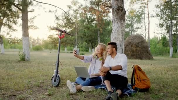 En medio de la naturaleza hermosa y carismática pareja tomar algunos selfies usando el teléfono inteligente que tienen un tiempo de picnic llegó con scooter eléctrico — Vídeos de Stock