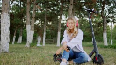 Güneşli bir günde, büyük gülüşlü sarışın bir kadın elektrikli scooterına biner ve telefona bakar ve kırmızı kulaklık takar.