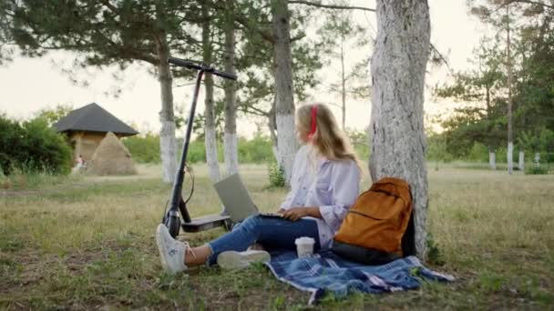 Na přírodu blond vlasy mladá žena s úžasným úsměvem si sednout na trávu drží notebook poslech hudby a zpěvu na sobě červené sluchátka — Stock video