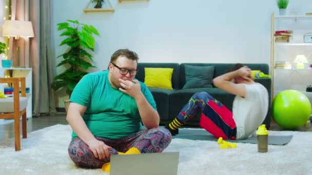 Вдома у вітальні здоровий хлопець їсть смачний бургер, в той час як його друг інший хлопець робить тренування, щоб отримати шість упаковок ожиріння хлопця, який дивиться фільм на ноутбук — стокове відео