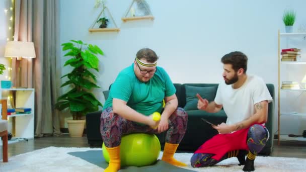 Charismatic obese guy and his friend at home doing the workout together a guy with a digital tablet helping the obese man to do the right exercises — Stock Video