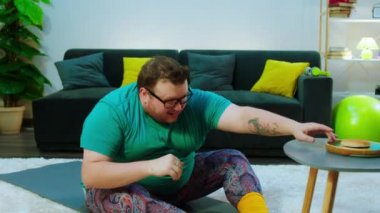 Obez komik adam evde hasırların üzerinde bir hamburger kokusu alıyor sonra da ağırlık kaldırmak için başka bir mekik çekiyor.