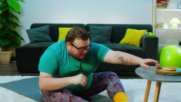 Übergewichtiger lustiger Kerl, der zu Hause auf der Matte Crunches macht, nimmt er einen Burger-Geruch und geht zurück, um weitere Crunches zu machen, um die Gewichte zu verlieren — Stockvideo