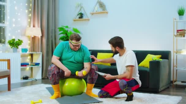Харизматичний чоловік допомагає своєму великому товстому другу робити тренування прямо вдома на підлозі, щоб схуднути — стокове відео