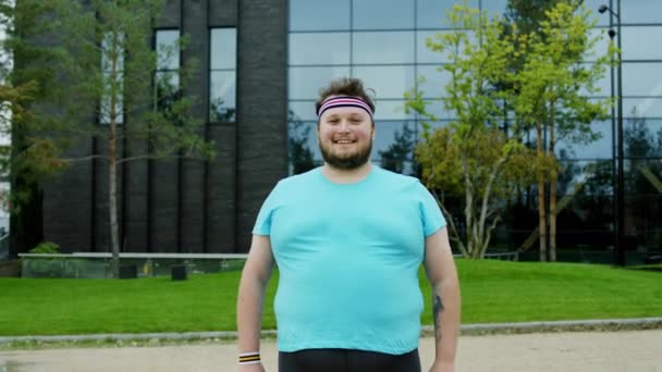 Usměje se velkému pózujícímu obéznímu mladíkovi před kamerou a připravuje se na aerobní cvičení venku vedle moderní budovy — Stock video