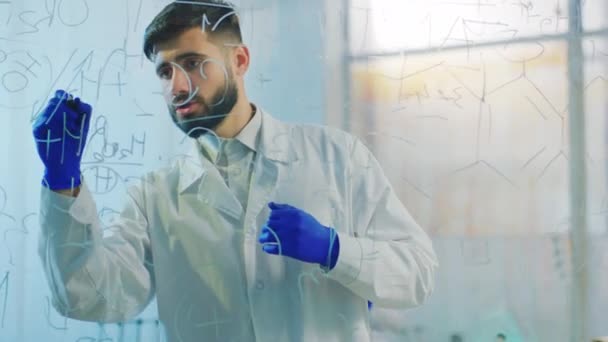 커다란 화학 실험실 과학자인 남자는 투명 한 판을 사용하여 얼마의 화학 공식을 작성하는데, 그의 동료 여자는 시험을 시작하기 전에 다른 화학 용액을 준비 한다. 4k — 비디오