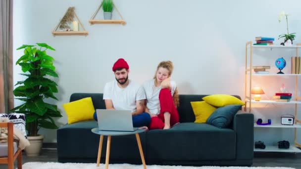 自宅でソファの上にかなりカップル一緒に楽しい時間を過ごす一緒にラップトップを使用して映画を見るために彼らはパジャマを着て — ストック動画