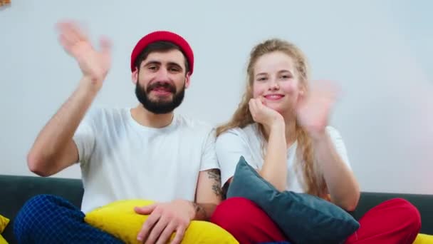 Vor der Kamera grüßt das Paar und lächelt groß in die Kamera, während es auf dem Sofa sitzt — Stockvideo