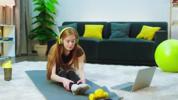 Eine Frau mit gelbem Headset sitzt auf der blauen Matte in einem hellen Raum, sie hat einen String-Kiefer und sieht sehr gut aus, sie ist auch fit, sie streckt ihren ganzen Körper zu ihren Zehen aus. — Stockvideo