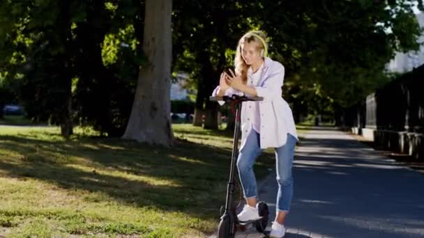 Vackra blonda hår dam med röda hörlurar mitt på gatan ta ett stopp med sin elektriska skoter för att sms:a någon. 4k — Stockvideo