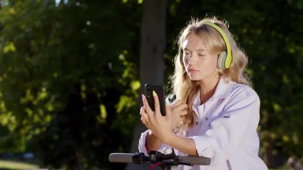 Na środku kamery skoncentrowana dama z długimi kręconymi włosami zatrzymuje się na środku ulicy ze swoim skuterem elektrycznym patrzy przez smartfona i dotyka swojej długiej blondynki — Wideo stockowe