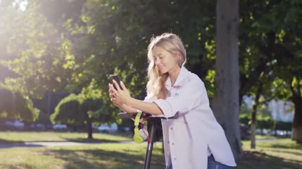 Ελκυστική ξανθιά κυρία μαλλιά στη μέση του δρόμου με ένα ηλεκτρικό σκούτερ κάνει μια στάση για να πάρετε μερικές selfies χρησιμοποιώντας το smartphone. 4ια — Αρχείο Βίντεο