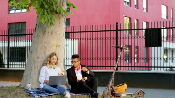 Dos trabajadores de oficina multirracial dama y chico en el almuerzo tomar un sentarse fuera en la hierba al lado de un gran árbol que utilizan el ordenador portátil para terminar un poco de trabajo — Vídeos de Stock