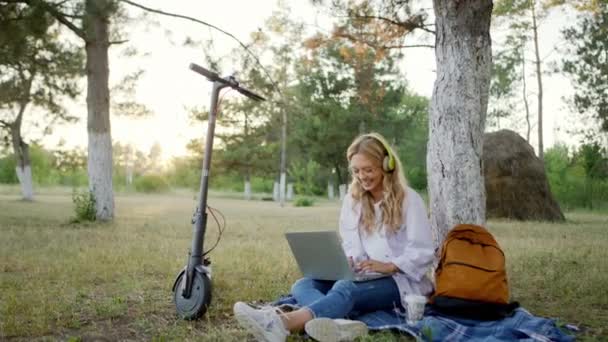 Emocionada estudiante llegó al parque con un scooter eléctrico tomar un sentarse en el césped y el uso de auriculares escuchando música al mismo tiempo haciendo su proyecto de la universidad en el ordenador portátil. Disparo en ARRI — Vídeo de stock