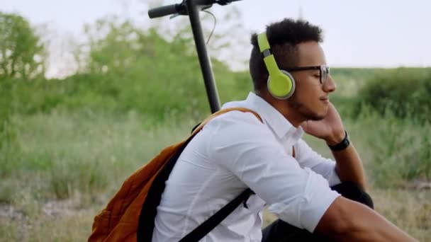 În fața camerei afro-american tip cu căști ascultând muzică în parc el ia un loc pe scuterul său electric și bucurându-se de momentul de la natură — Videoclip de stoc