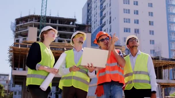 Foreman e outros especialistas em construção diversos usando laptop ração que trabalham no meio do canteiro de obras usando uma ração para discutir com outros trabalhadores da construção — Vídeo de Stock