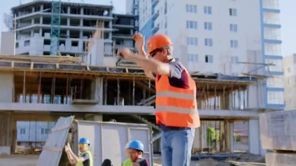 Před kameramanem předák středního věku tančí a skáká v době oběda si užívá čas se svým týmem na staveništi má na sobě bezpečnostní vybavení — Stock video