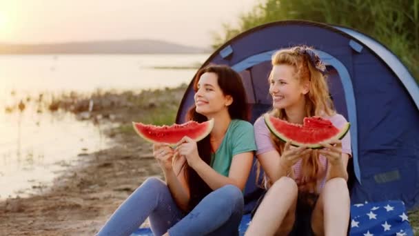 Hermosas mujeres jóvenes al atardecer junto al lago y la tienda de campaña tienen un tiempo de picnic que comer algunos trozos de sandía y sentirse relajado. Disparo en ARRI Alexa — Vídeo de stock