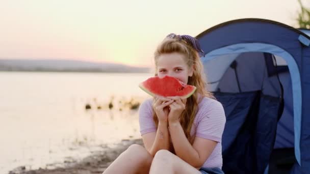 Перед камерой красивая леди с идеальной улыбкой на пикнике на закате у озера, поедающая перед камерой кусок арбуза — стоковое видео