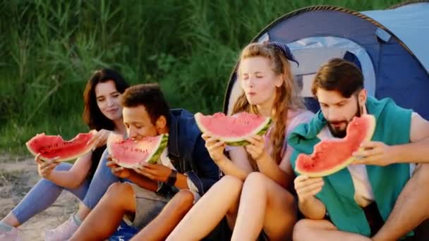 Al atardecer buen aspecto grupo diverso de amigos comiendo deliciosos trozos de sandía junto al lago en el picnic al atardecer — Vídeo de stock